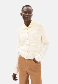 Krämfarbener gestrickter Cardigan mit horizontalen Mustern und Frontknöpfen, kombiniert mit einem hellgelben Hemd mit Kragen und braunen Cordhosen.