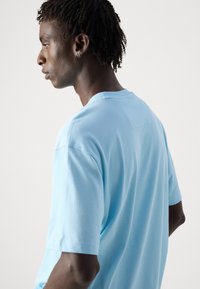 Ανοιχτό μπλε βαμβακερό μπλουζάκι με στρογγυλή λαιμόκοψη, κοντά μανίκια και ελάχιστες ραφές, απεικονιζόμενο από πλάγια και πίσω.