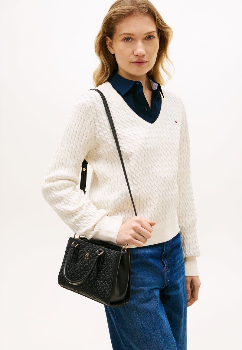 Γυναίκα με ξανθά μαλλιά που φορεί λευκό πουλόβερ πλεκτό με κοτσίδες, μπλε πουκάμισο με γιακά, μπλε τζιν, κρατώντας μαύρη τσάντα με σχέδια και ιμάντα ώμου.