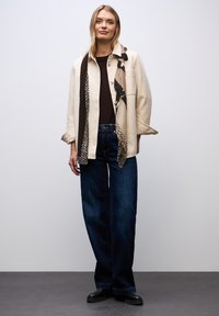 Beige jakke med brede ærmer, sort top, mørkeblå jeans og et mønstret tørklæde med brune, beige og sorte detaljer. Glat tekstur, afslappet pasform.