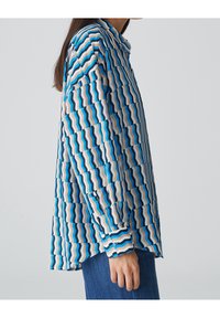 Koszula z długim rękawem, z zygzakowatym wzorem w odcieniach niebieskiego, beżu i brązu. Wykonana z gładkiego materiału, o luźnym kroju, z detalami w postaci guzików.