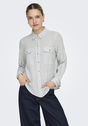 Mujer vistiendo una camisa de manga larga con botones, a rayas verticales blancas y grises, y vaqueros azul oscuro de pierna ancha.