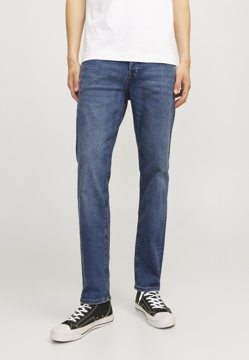 Jeans de mezclilla azul con corte recto, que presentan un ligero desgaste, diseño de cinco bolsillos y cierre de botón, combinados con zapatillas negras.