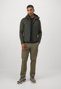 Gewatteerde olijfgroene vest met een hoge kraag, gecombineerd met een zwarte longsleeve shirt en licht olijfgroene cargo broek, afgewerkt met wandelschoenen.