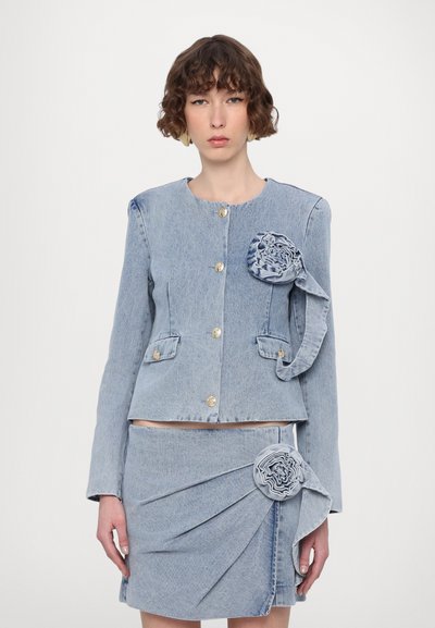 Veste en denim bleu clair avec des boutons dorés, dotée d'un détail de tissu floral sur l'épaule. Associée à une jupe assortie avec des plis.