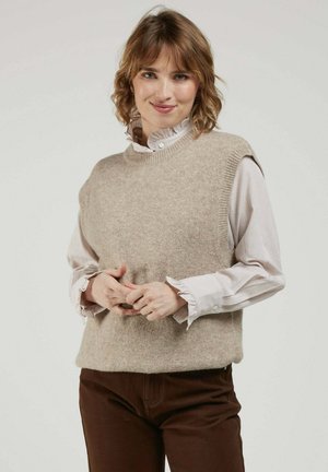 Femme aux cheveux courts et ondulés portant un pull sans manches beige par-dessus un chemisier blanc à volants et un pantalon marron, debout devant un fond uni.