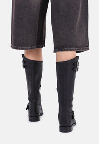 Botas altas de cuero negro con una textura suave, que presentan dos detalles de hebilla y una cremallera lateral, ubicadas sobre un fondo neutral.