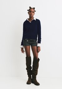 Marineblauer Strickpullover mit weißem Kragen, graue Shorts und braune, lässige Oberschenkelstiefel. Verfügt über einen doppelten Schnallen-Gürtel.