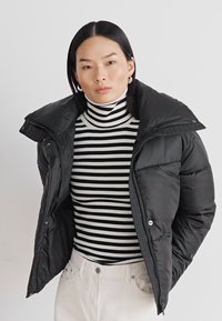 Svart pufferjakke med høy krage og trykknapper, båret over en svart og hvit stripete high-neck genser, kombinert med lyse bukser.