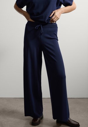 Personne portant un pantalon large bleu marine avec une taille à cordon et des mocassins marron foncé, se tenant les mains près de la taille contre un mur uni.