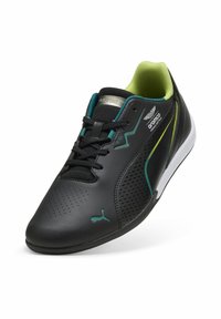 Svart träningsskor med perforerat mönster, gröna accenter och vit sula. Har svarta snören och en strukturerad yta. Logotyp på sidan.