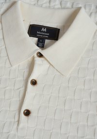 Tejkrém színű pólóing, textúrált kockás mintával, gallérral, három barna gombbal és a márkát azonosító címkével.