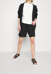 Svart zip-up hoodie, vit Nike t-shirt, svarta shorts, svarta sandaler med vit plattform, person som står på en enfärgad bakgrund.