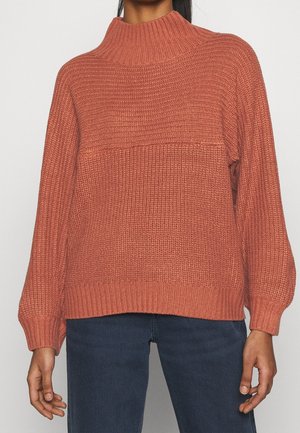 Frau trägt einen rostfarbenen, texturierten Strickpullover mit hohem Rundhalsausschnitt und langen Ballonärmeln, kombiniert mit dunkelblauen Jeans.