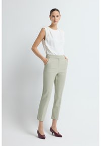 Vrouw met een knot in haar haar, gekleed in een mouwloos wit topje, lichtgrijze cropped broek en bordeauxrode hakken, staande voor een witte achtergrond.