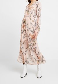 Jurk met lange mouwen en V-hals van zachtroze stof met bloemenpatronen, ruches aan de zoom en doorschijnende textuur, gecombineerd met witte enkellaarsjes.