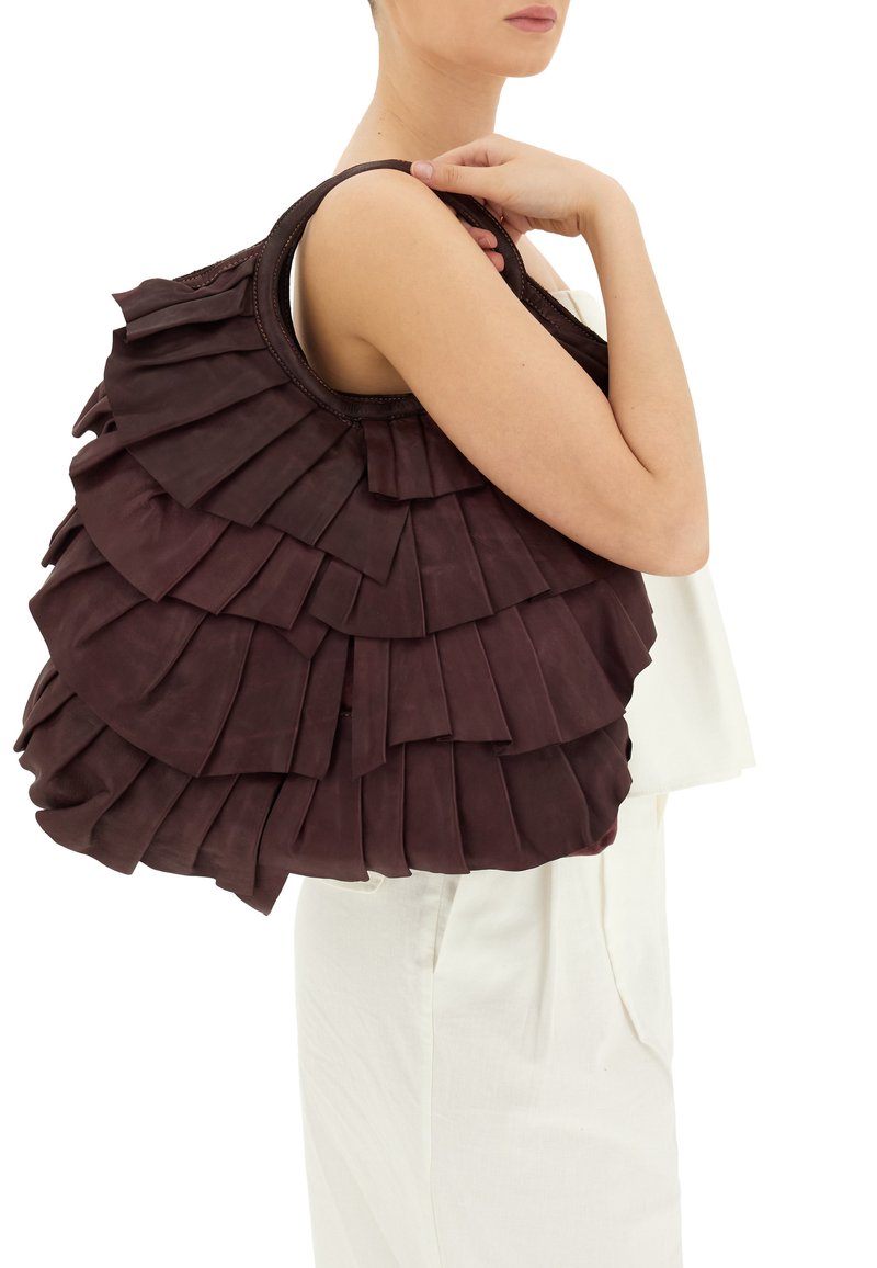 Borsa in pelle bordeaux con design a strati e volant. Texture morbida e manico arrotondato. Modello che tiene la borsa contro un outfit chiaro.