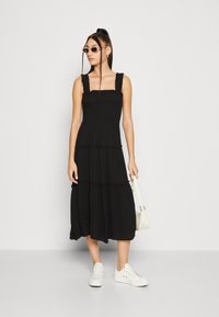 Robe maxi noire à volants avec décolleté carré, bretelles froncées et tissu fluide. Portée avec des baskets blanches et un petit sac blanc.