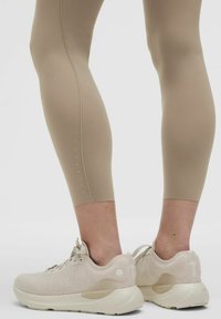 Könnyű bézs leggings sima, rugalmas anyagból, oldalvarrással és kis szellőzőnyílásokkal. Világos bézs sportcipők texturált szövettel és párnázott talppal.
