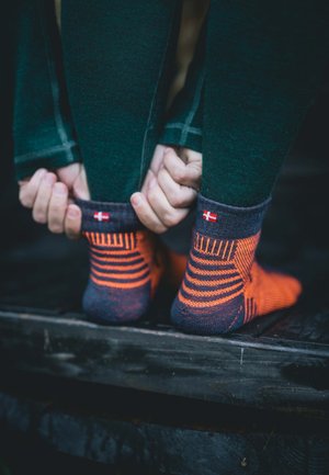 Ankelstrømper med en mørkegrå bund, orange striber og en lille flagdetalje. Materialet ser tekstureret og strækbart ud.