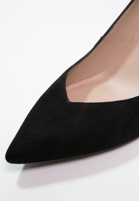 Chaussure plate en daim noir à bout pointu, empeigne en V et doublure intérieure nude sur fond blanc.