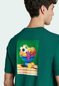 Camiseta de algodón verde con un gráfico colorido en la parte trasera de un tazón con frutas y pelotas deportivas, incluyendo naranjas y una pelota de fútbol.