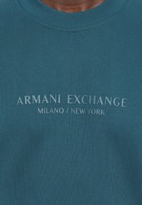 Teal langermet crewneck skjorte laget av teksturert stoff med et hevet logodesign: "ARMANI EXCHANGE MILANO / NEW YORK" i lys farge.
