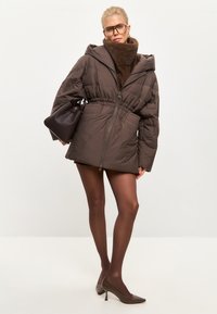 Bruine puffer jas met een capuchon, een getailleerde taille en een rits. Draag met een donkerbruine sjaal, doorschijnende panty's en bruin puntige hakken. Bruine handtas.