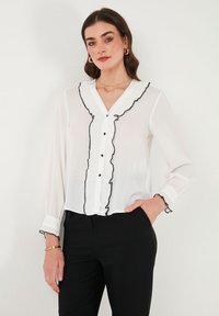 White blouse with black ruffled trim, button-down front, long sleeves with elastic cuffs, lightweight fabric, paired with black pants.