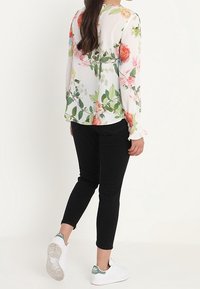 Blouse à fleurs à manches longues blanche, ornée de roses colorées et de feuilles ; associée à un pantalon noir et des baskets blanches avec des accents verts.