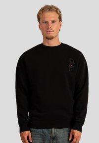 Felpa nera realizzata in tessuto spesso con colletto rotondo, caratterizzata da una figura di cartone ricamata con un cuore sul lato sinistro del petto.