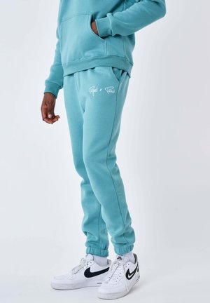 Persona che indossa pantaloni della tuta turchese e felpa abbinata con scritta "Project x Paris", abbinati a sneaker bianche Nike Air.