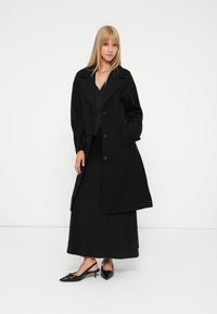 Casaco de lã preto com gola em bico, botões duplos à frente e bolsos laterais. Combinado com um vestido preto e sapatos de bico fino.