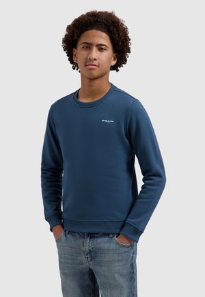 Jong persoon met krullend haar, draagt een blauwe trui met het logo "BALLIN" en lichtblauwe jeans, handen in de zakken, neutrale achtergrond.