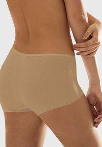 Frau in beiger nahtloser Unterwäsche, von der unteren Rückseite und den Hüften gezeigt, mit einer Hand, die ihren Oberarm berührt.