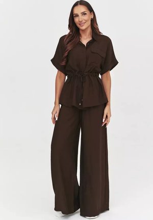 Kobieta o długich brązowych włosach, ubrana w ciemnobrązowy krótki rękaw z guzikami z wciętą talią i szerokimi spodniami, stojąca i uśmiechająca się.