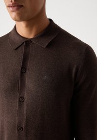 Bruine gebreide trui met een kraag, knoopsluiting en een subtiele geborduurde logo op de borst. Zacht, textuurgegeven stof.