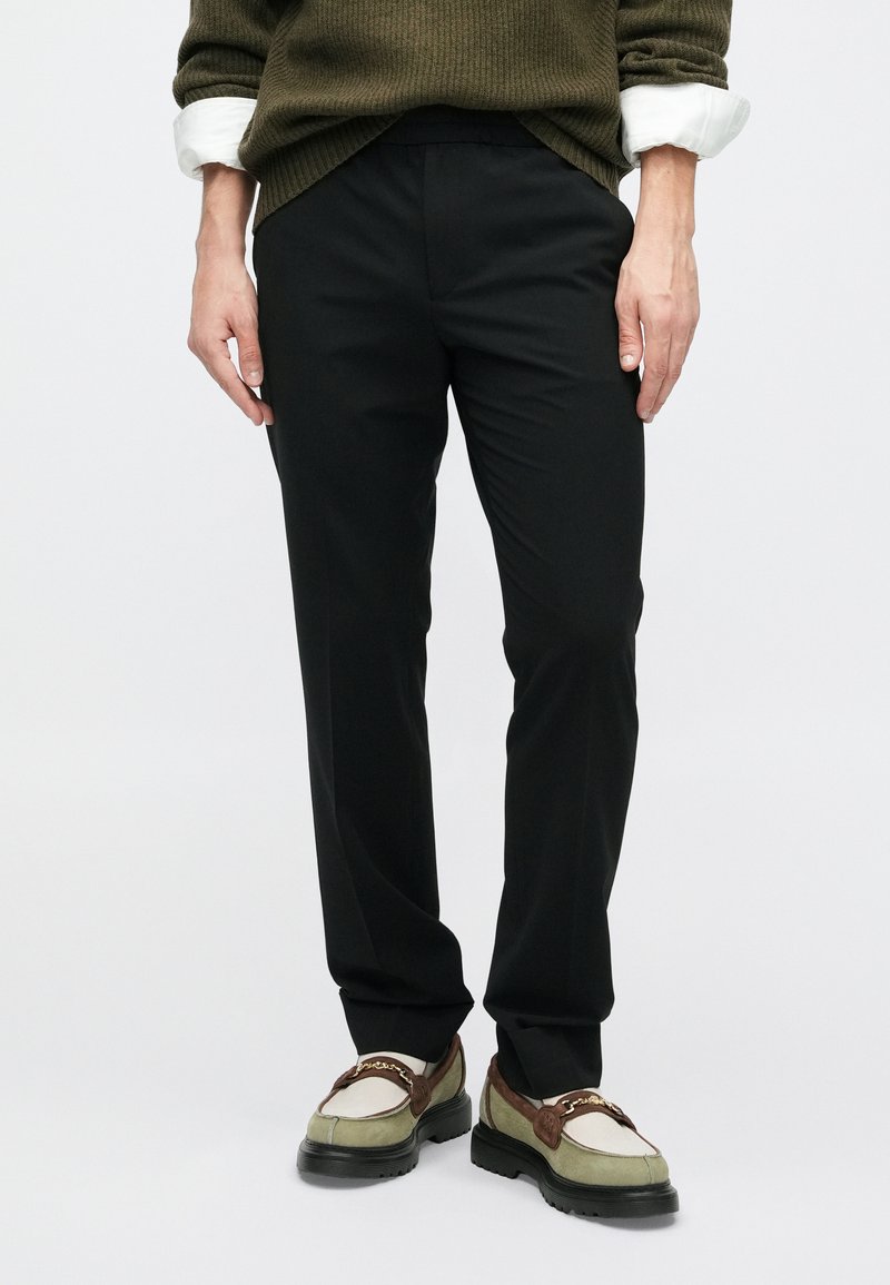 Zwarte op maat gemaakte broek met een gladde afwerking, met een eenvoudig ontwerp en een rechte pasvorm. Gecombineerd met groene en bruine loafers.