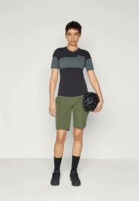 Majica s kratkimi rokavi v črno in teal prugah s sprednjo žepom, v kombinaciji z olivno zelenimi kratkimi hlačami. Model drži črn čelado za kolo.