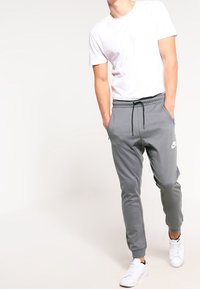 Man i vit t-shirt och grå Nike sweatpants med händerna i fickorna, iförd vita sneakers, stående framför en ljus bakgrund.