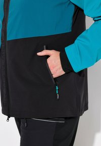 Veste imperméable en teal et noir. Comprend une poche latérale zippée avec une quincaillerie bleue et un tissu texturé. Design moderne et ajusté.