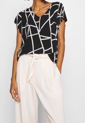 Mujer con camiseta negra de manga corta con patrón abstracto de rejilla blanca y pantalones blancos de cintura alta con cinturón atado.
