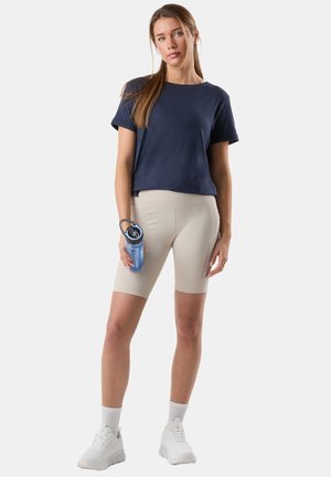 Young woman standing, wearing navy t-shirt, beige bike shorts, white sneakers, holding blue water bottle with black lid against white background.