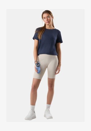 Young woman standing, wearing navy t-shirt, beige bike shorts, white sneakers, holding blue water bottle with black lid against white background.