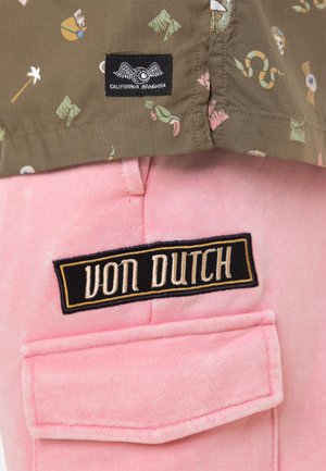 Gros plan de l'ourlet d'une chemise kaki avec de petites impressions colorées au-dessus d'une poche en tissu rose étiquetée "VON DUTCH" sur un patch noir avec broderie beige.
