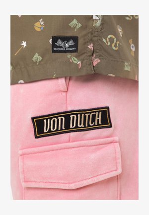 Gros plan de l'ourlet d'une chemise kaki avec de petites impressions colorées au-dessus d'une poche en tissu rose étiquetée "VON DUTCH" sur un patch noir avec broderie beige.