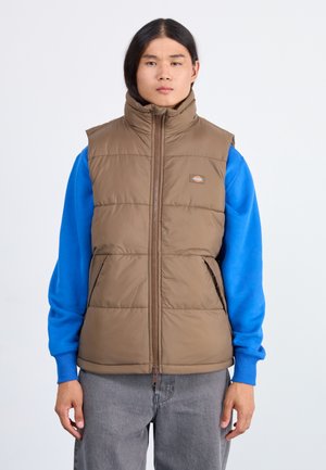 Brown padded vest with high collar and front zipper. Worn over a blue long-sleeve shirt. Features side pockets and logo patch on chest.