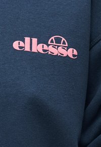 Σκούρο μπλε ύφασμα με ροζ λογότυπο "ellesse" που περιλαμβάνει γραφικό με ημικύκλιο τένις πάνω από το κείμενο.