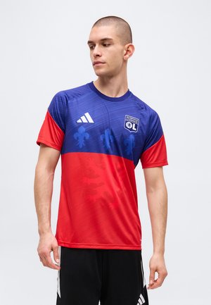 Muž v modro-červenom futbalovom drese Olympique Lyonnais s logami tímu a Adidas, pozerá doľava na jednofarebnom pozadí.