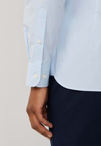 Svijetloplava košulja dugih rukava s oštrim reverima, manžetama na jedan gumb i glatkom teksturom, kombinirana s tamnoplavim materijalom ispod.